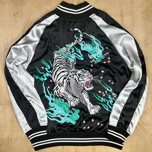 Puede incluir: Chaqueta bomber de satén negro con un bordado de tigre blanco en la espalda. La chaqueta presenta mangas plateadas y ribete a rayas blanco y negro en el cuello, los puños y el dobladillo. El tigre está representado con la boca abierta, rodeado de detalles turquesa y rosa.