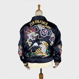 Könnte beinhalten: Eine schwarze Satin-Bomberjacke mit aufwendiger Rückenstickerei eines Drachen, eines Tigers, eines Berges und Wolken. Der Schriftzug OKINAWA ist in Gelb über dem Drachen gestickt. Zusätzliche Drachenstickereien befinden sich an den Ärmeln.