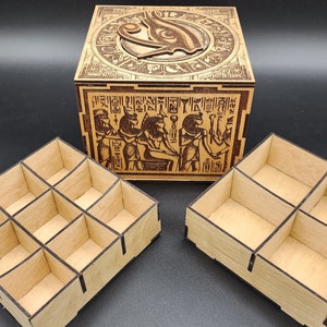 Puede incluir: Una caja de madera con un diseño intrincado de jeroglíficos egipcios y un ojo de Horus tallado. La caja tiene dos bandejas extraíbles con compartimentos para organizar objetos pequeños.