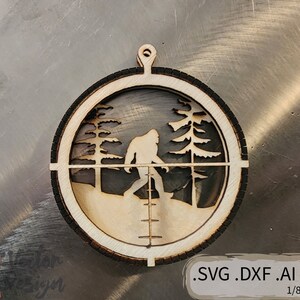 May include: A wooden Christmas ornament featuring a silhouette of Bigfoot walking through a forest. The ornament is in the shape of a circle with a crosshair design. The text .SVG .DXF .AI .PDF 1/8" (3MM) is printed below the ornament.