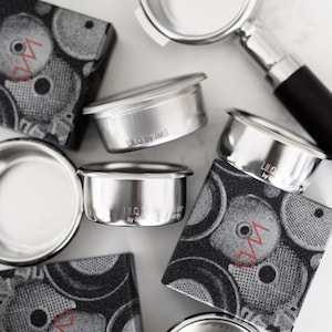 Puede incluir: Un juego de cinco portafiltros de espresso plateados con el texto "LILO by IMS" grabado en el lateral. Los portafiltros están sobre una superficie con estampado en blanco y negro.