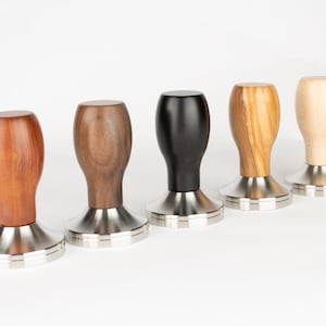 May include: Five espresso tampers with different colored wooden handles. The tampers have a silver base and a round wooden handle. The handles are brown, dark brown, black, light brown, and white.