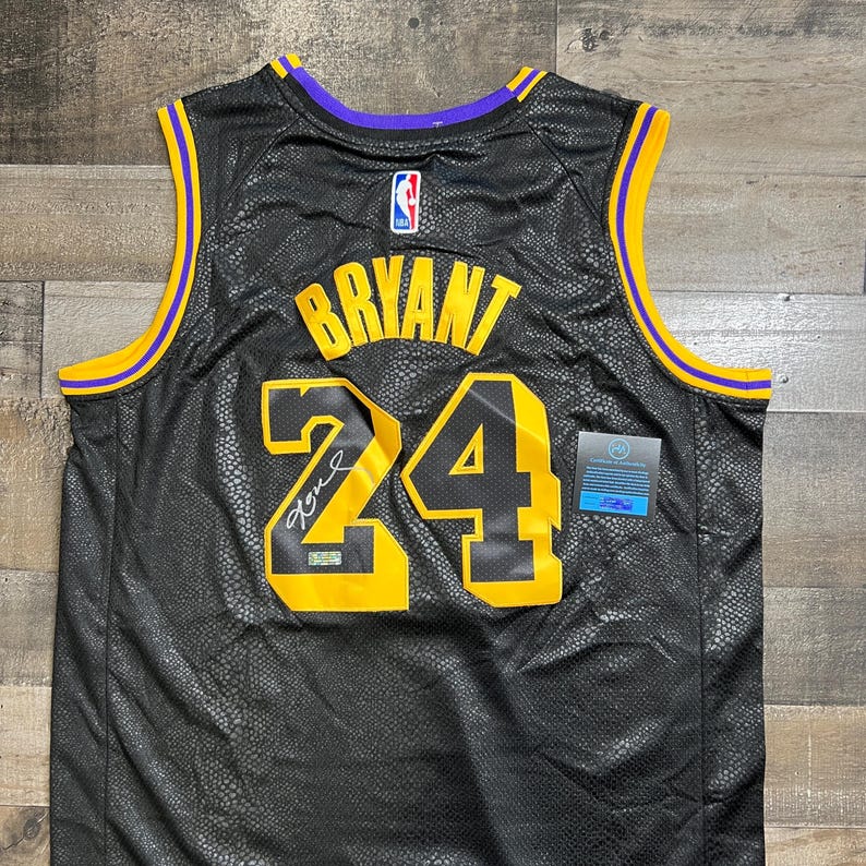 May include: A black basketball jersey with a snakeskin-like texture, featuring the name BRYANT and the number 24 in yellow with black outlines. The jersey has purple and yellow trim on the collar and armholes, an NBA logo, and a signature on the number.