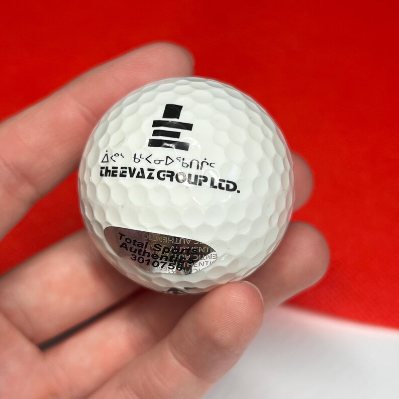 May include: A white golf ball with a dimpled surface. The ball has black text and a logo. The text "CHEEVAZ GROUP LTD." is printed on the ball. A silver sticker with text is also on the ball.