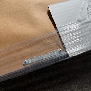 Puede incluir: La parte posterior de un objeto enmarcado con un fondo de papel marr&oacute;n y un protector de esquina de cart&oacute;n blanco. Un colgador de sierra de metal plateado est&aacute; fijado con dos tornillos al marco, todo envuelto en film pl&aacute;stico transparente.