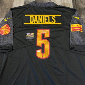 May include: A black sports jersey featuring the name DANIELS and the number 5 in yellow with red outlines. The sleeves have a grey honeycomb pattern, a yellow Nike logo, a Commanders patch, and a D.C. flag patch. An authentication sticker is present.