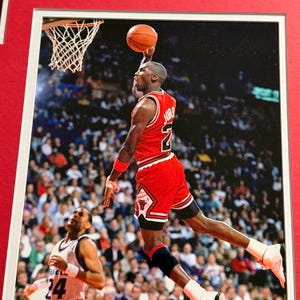 Puede incluir: Una fotograf&iacute;a enmarcada de un jugador de baloncesto con el uniforme rojo y blanco de los Chicago Bulls con el nombre JORDAN y el n&uacute;mero 23 en la espalda, capturado en el aire mientras encesta un bal&oacute;n naranja durante un partido profesional.