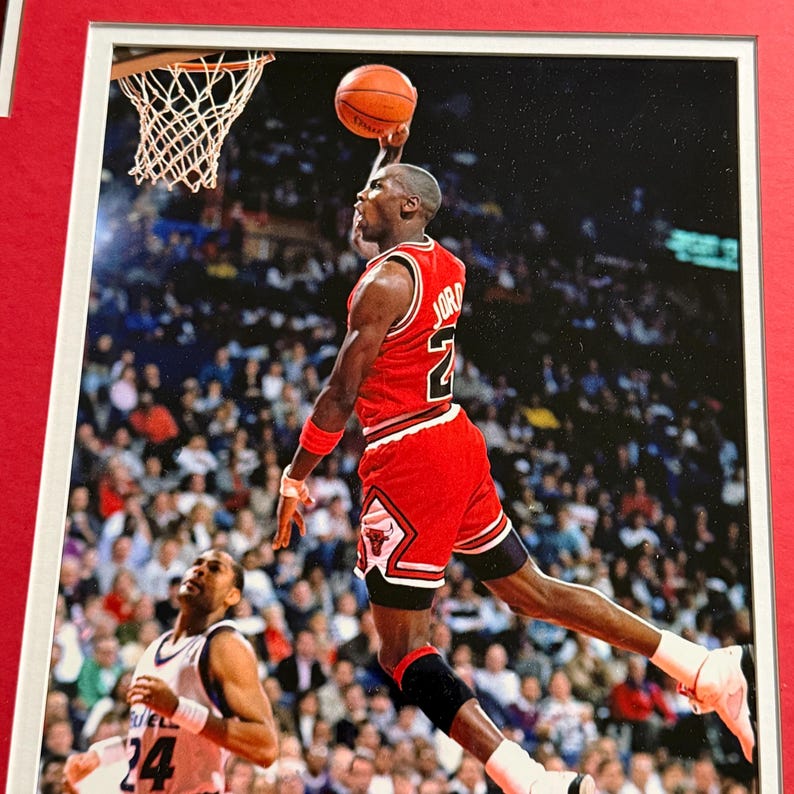 Puede incluir: Una fotograf&iacute;a enmarcada de un jugador de baloncesto con el uniforme rojo y blanco de los Chicago Bulls con el nombre JORDAN y el n&uacute;mero 23 en la espalda, capturado en el aire mientras encesta un bal&oacute;n naranja durante un partido profesional.