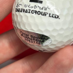 May include: A white golf ball with a dimpled surface. The ball has black text that reads "THE EVAZ GROUP LTD." and a sticker with additional text. The ball is held by a person.