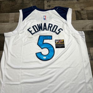 Puede incluir: Una camiseta de baloncesto blanca con paneles de hombro azul oscuro y el nombre EDWARDS en letras azul oscuro sobre el número 5 en azul claro con un borde azul oscuro. La camiseta incluye un logotipo de la NBA y una pequeña etiqueta de autenticidad.