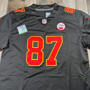 May include: A black football jersey with the number 87 in red and gold. The jersey has red and gold accents on the shoulders and a team logo patch on the chest. The NFL logo is visible on the neckline.