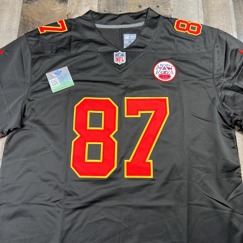 May include: A black football jersey with the number 87 in red and gold. The jersey has red and gold accents on the shoulders and a team logo patch on the chest. The NFL logo is visible on the neckline.