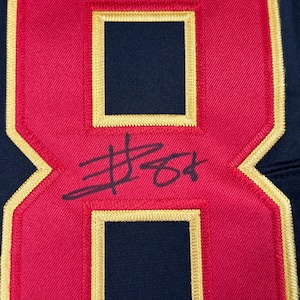 May include: Close-up of a red and navy blue jersey number 8 with gold trim. The number has a signature in black ink. The jersey appears to be made of a textured fabric.