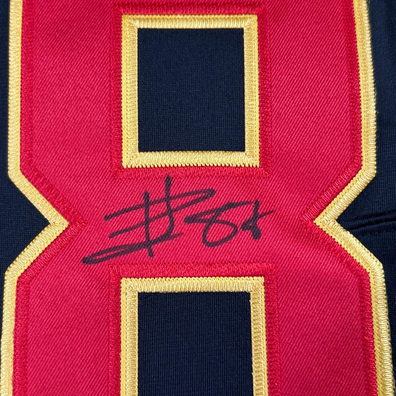 May include: Close-up of a red and navy blue jersey number 8 with gold trim. The number has a signature in black ink. The jersey appears to be made of a textured fabric.