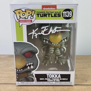 May include: A Tokka vinyl figure from the Teenage Mutant Ninja Turtles Movies series. The figure is in a clear plastic display case. The figure is green, gray, and yellow with red eyes. The box has the number 1139.