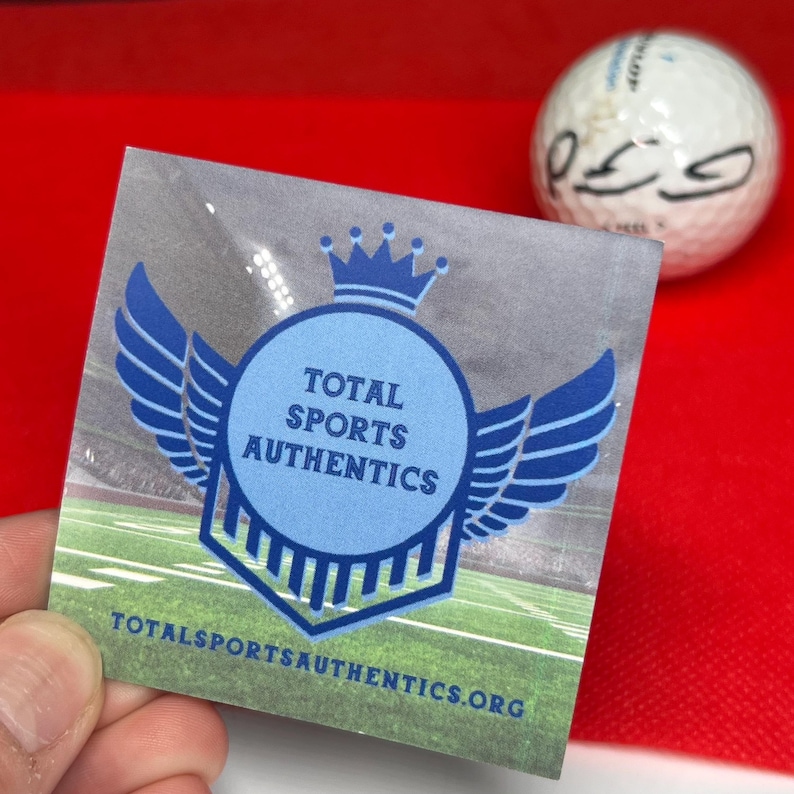 May include: A clear sticker with the text "TOTAL SPORTS AUTHENTICS" in a blue circle with wings and a crown. A white golf ball with black markings is in the background. The sticker also has the website address: TOTALSPORTSAUTHENTICS.ORG.