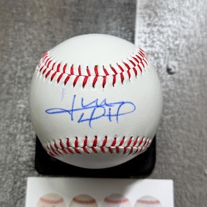 Op de afbeelding: Een witte honkbal met rode stiksels en een handtekening in blauwe inkt in het midden. De bal staat op een zwarte standaard, met een kleine witte kaart met afbeeldingen van honkballen zichtbaar aan de onderkant van het beeld.
