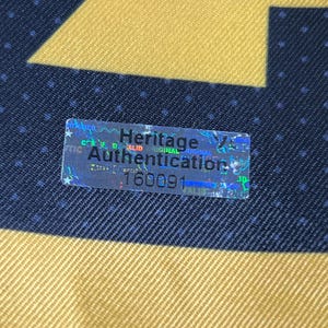 May include: A rectangular holographic authentication sticker with a silver finish, featuring the black text Heritage Authentication and the number 160091, placed on a dark blue fabric background with a gold geometric pattern and small white dots.