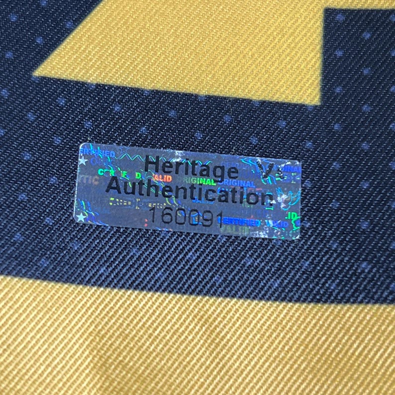 May include: A rectangular holographic authentication sticker with a silver finish, featuring the black text Heritage Authentication and the number 160091, placed on a dark blue fabric background with a gold geometric pattern and small white dots.