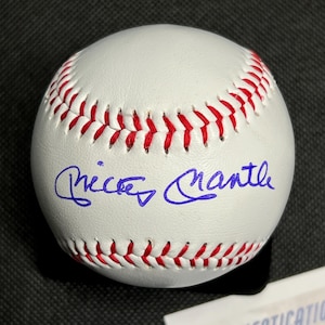 May include: A white baseball with red stitching and a blue signature. The baseball is likely an autographed collectible. The signature appears to read "Mickey Mantle". The ball is on a dark surface.