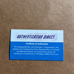 Puede incluir: Una tarjeta rectangular de certificado de autenticidad de Authentication Direct. La secci&oacute;n superior es blanca con el texto AUTHENTICATION DIRECT en azul oscuro. La secci&oacute;n inferior es azul con texto blanco que detalla el certificado.