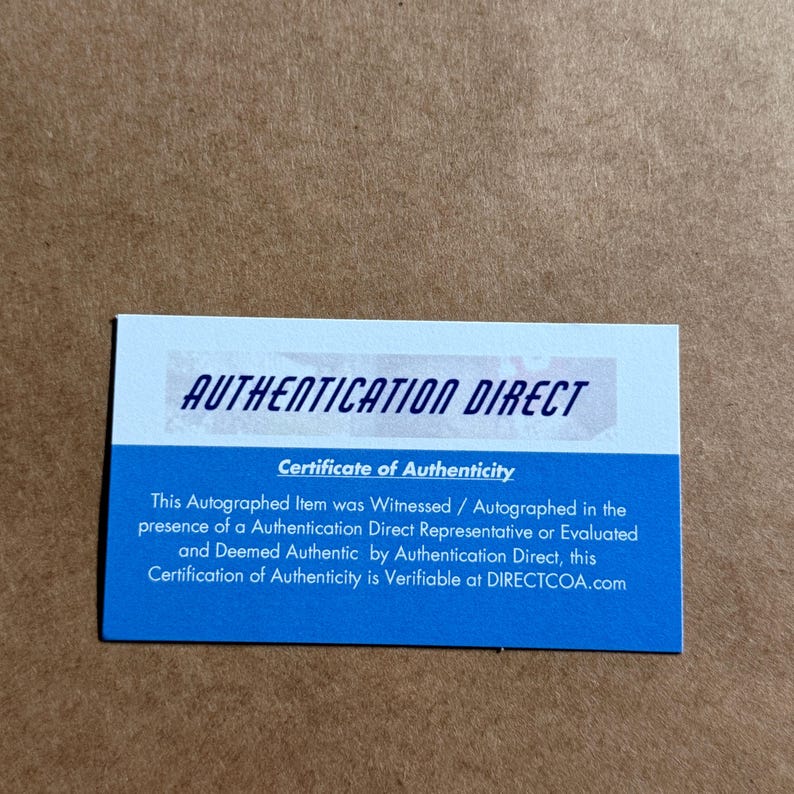 Puede incluir: Una tarjeta rectangular de certificado de autenticidad de Authentication Direct. La secci&oacute;n superior es blanca con el texto AUTHENTICATION DIRECT en azul oscuro. La secci&oacute;n inferior es azul con texto blanco que detalla el certificado.