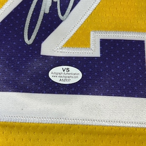 May include: Close-up of a yellow and purple sports jersey with white trim. The jersey has a large number and a VS Autograph Authentication sticker. The sticker includes the website www.vsautographs.com and the number A52537.
