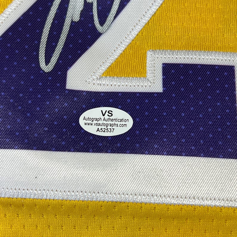 May include: Close-up of a yellow and purple sports jersey with white trim. The jersey has a large number and a VS Autograph Authentication sticker. The sticker includes the website www.vsautographs.com and the number A52537.