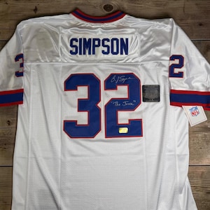 May include: A white sports jersey featuring the name SIMPSON and the number 32 in blue with red trim on the back. The jersey includes a blue and red collar, blue and red sleeve cuffs, an autograph with the inscription The Juice, and an NFL tag.