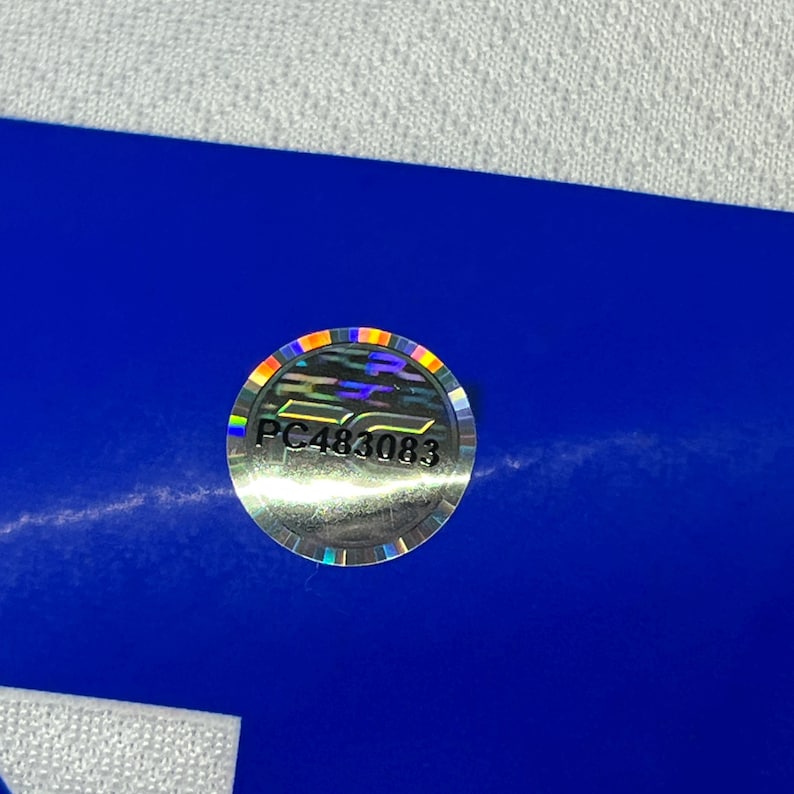 Puede incluir: Una pegatina hologr&aacute;fica redonda con el texto "PC483083" en plata. La pegatina tiene un borde de color arco&iacute;ris y est&aacute; colocada sobre un fondo azul. Es probable que sea una etiqueta o sello de producto.