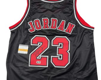 Puede incluir: Una camiseta de baloncesto negra con el nombre JORDAN y el número 23 en rojo con bordes blancos. La camiseta tiene ribetes rojos y blancos en el cuello y las sisas, una textura de malla y una firma sobre el número 23 con una etiqueta.