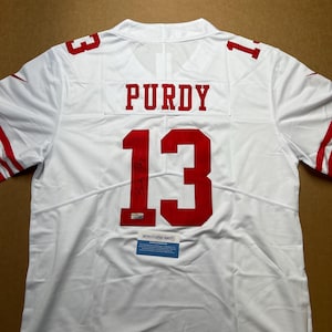 May include: A white football jersey featuring the name PURDY and the number 13 in red on the back. The jersey includes a signature on the number 13, a small authentication sticker, and a rectangular Authentication Direct certificate of authenticity.
