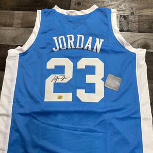 K&ouml;nnte beinhalten: Hellblaues Basketballtrikot mit dem Namen "JORDAN" und der Nummer "23" in Wei&szlig;. Das Trikot hat einen wei&szlig;en Kragen und wei&szlig;e Seitenteile. Enth&auml;lt eine Unterschrift und einen kleinen goldenen Aufkleber.