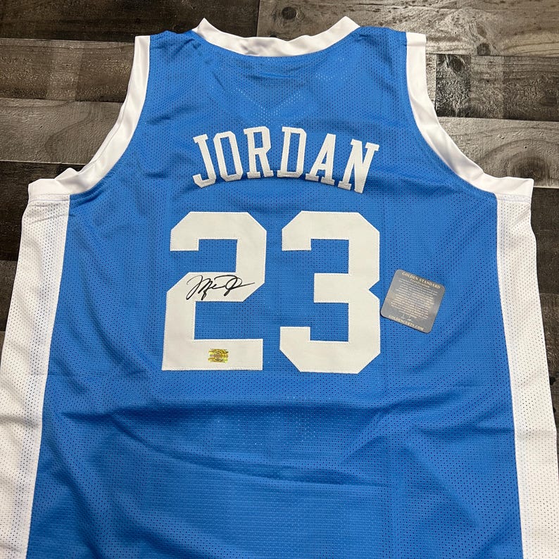 K&ouml;nnte beinhalten: Hellblaues Basketballtrikot mit dem Namen "JORDAN" und der Nummer "23" in Wei&szlig;. Das Trikot hat einen wei&szlig;en Kragen und wei&szlig;e Seitenteile. Enth&auml;lt eine Unterschrift und einen kleinen goldenen Aufkleber.