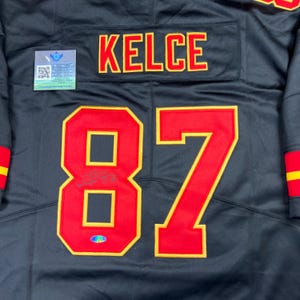 May include: Black jersey with the name "KELCE" in red letters with yellow trim across the upper back. The number "87" in red with yellow trim is on the back. A small tag with a QR code is on the upper left.