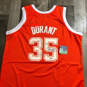 May include: An orange basketball jersey with white trim around the collar and armholes. The name "DURANT" is across the top, with the number "35" below. A signature and a small tag are visible on the jersey.