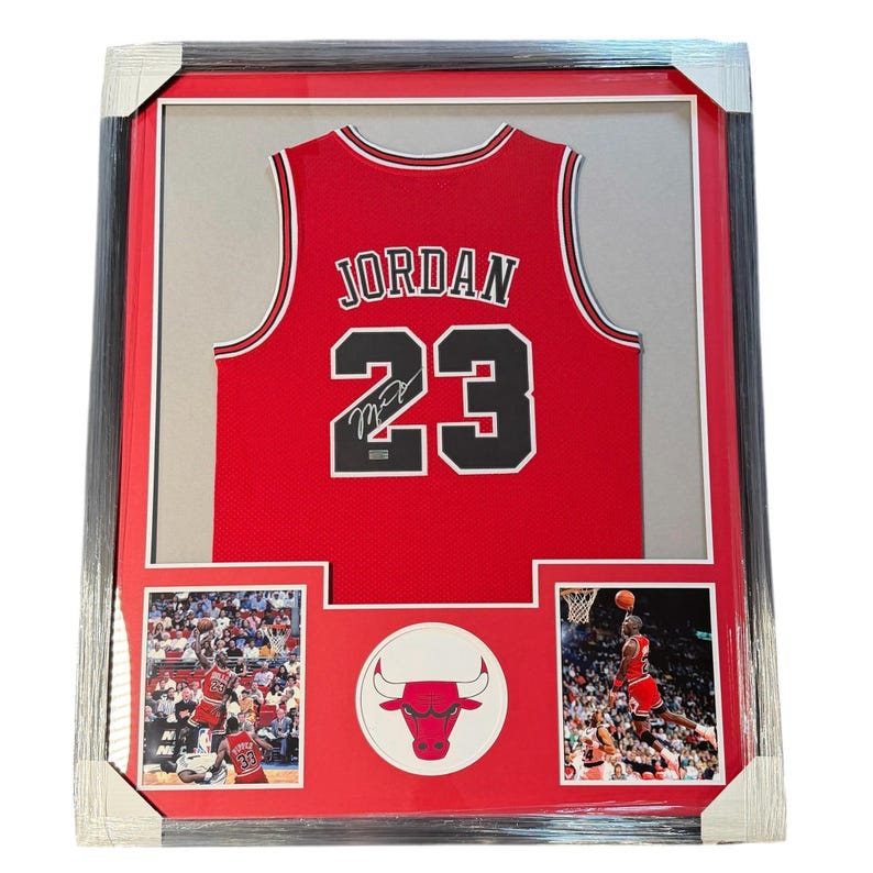 Puede incluir: Una camiseta de baloncesto roja de los Chicago Bulls enmarcada con el nombre JORDAN y el n&uacute;mero 23 en letras negras y blancas. La camiseta presenta una firma sobre el n&uacute;mero 23, acompa&ntilde;ada de dos fotos de acci&oacute;n y un emblema circular de los Bulls.