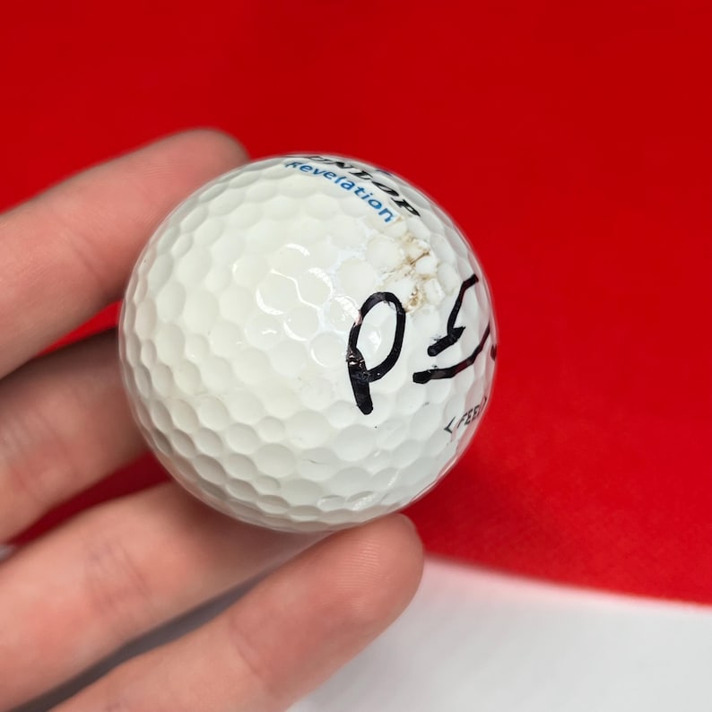 May include: A white golf ball with a dimpled surface, featuring black handwritten markings. The ball has a small scuff mark and the text "Dunlop Revelation" is visible. The background is a solid red.