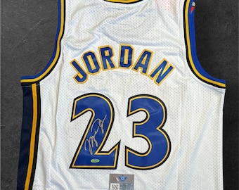 May include: White basketball jersey with blue and gold trim. The name "JORDAN" is across the shoulders in gold lettering. The number "23" is displayed in blue with a signature below. A small authentication sticker is visible.