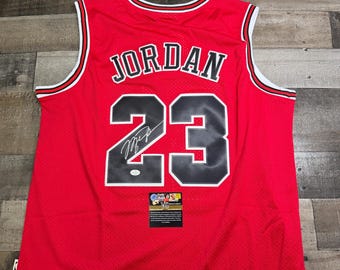 May include: A red basketball jersey featuring the name JORDAN and the number 23 in black with white outlines. The jersey has white and black trim on the collar and armholes. A signature is visible on the number 2, and a small authentication label is attached.