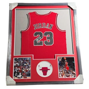 Puede incluir: Una camiseta de los Chicago Bulls enmarcada con el nombre JORDAN y el número 23 en negro con borde blanco. La camiseta incluye una firma, dos fotografías de acción de un jugador de baloncesto y un emblema central circular del logo Bulls.