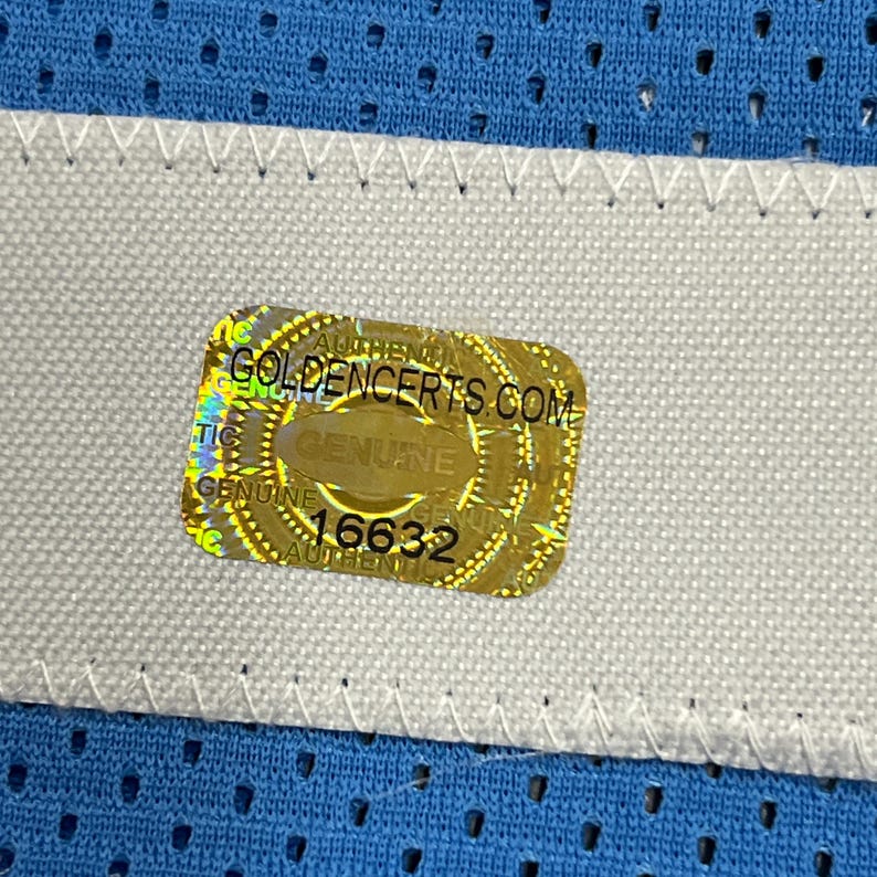 K&ouml;nnte beinhalten: Nahaufnahme eines blauen Mesh-Stoffs mit einem wei&szlig;en Streifen und einem goldenen Aufkleber. Der Aufkleber lautet "GOLDENCERTS.COM" und "GENUINE" mit der Nummer "16632". Der Stoff hat ein strukturiertes Aussehen.