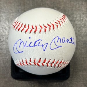 May include: A white baseball with red stitching and a blue signature that reads "Mickey Mantle." The baseball is resting on a black display stand. A collectible sports item.