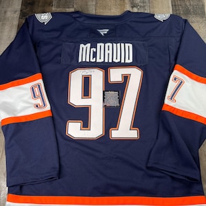 May include: A navy blue hockey jersey featuring the name "McDAVID" and the number "97". The jersey has white and orange detailing on the sleeves and hem. The number "9" is on the left sleeve and "7" on the right sleeve. The jersey is a sports item.