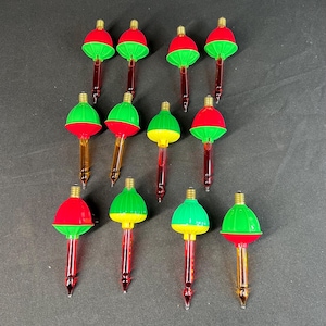 May include: A collection of vintage Christmas light bulbs. The bulbs have a red and green color scheme, with a clear, pointed base. The bulbs are arranged on a dark surface.