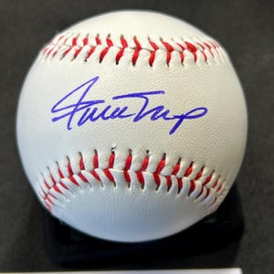 May include: A white baseball with red stitching and a blue signature. The baseball is resting on a black surface. The signature is in blue ink.