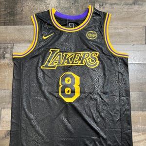May include: A black sleeveless basketball jersey with a snakeskin-like texture. It features the yellow and purple Lakers logo, the number 8 in yellow and black, a yellow Nike swoosh, and a circular Bibigo logo on the chest with purple and yellow trim.