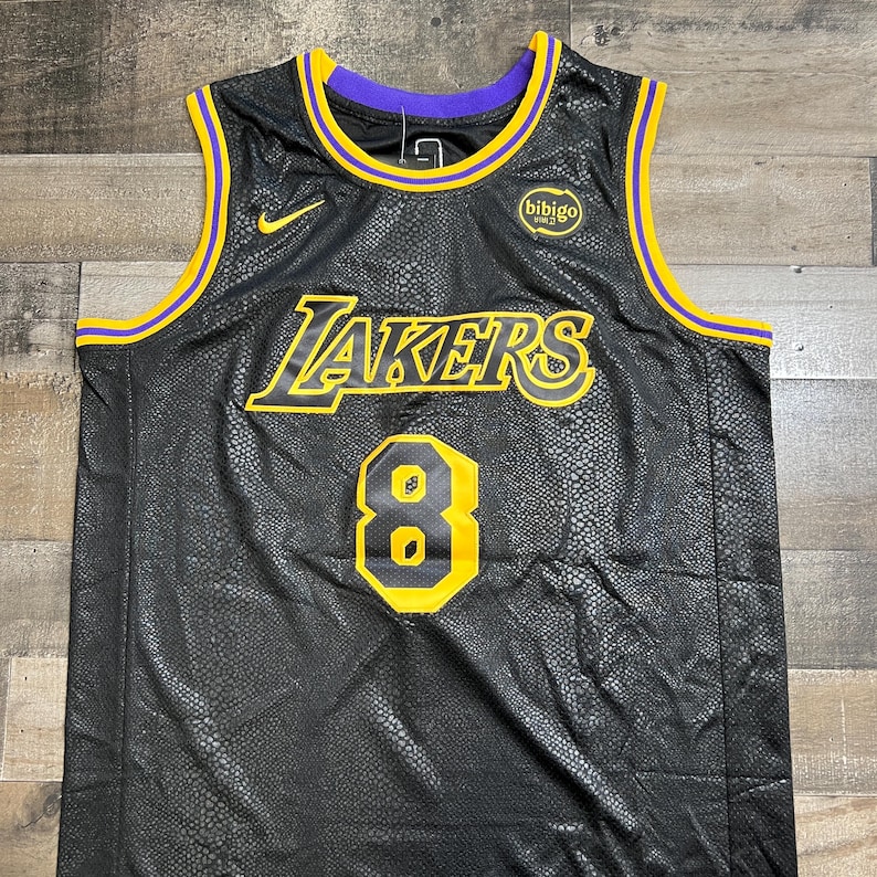 May include: A black sleeveless basketball jersey with a snakeskin-like texture. It features the yellow and purple Lakers logo, the number 8 in yellow and black, a yellow Nike swoosh, and a circular Bibigo logo on the chest with purple and yellow trim.