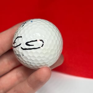 May include: A white golf ball with a dimpled surface. The ball has black markings and the word "FEEL" printed on it. The background is red and white.