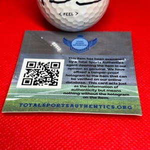 May include: A white golf ball with the word "< FEEL >" printed on it, resting above a rectangular card. The card features a QR code, a logo, and text that reads "TOTAL SPORTS AUTHENTICS". The card is on a red surface.